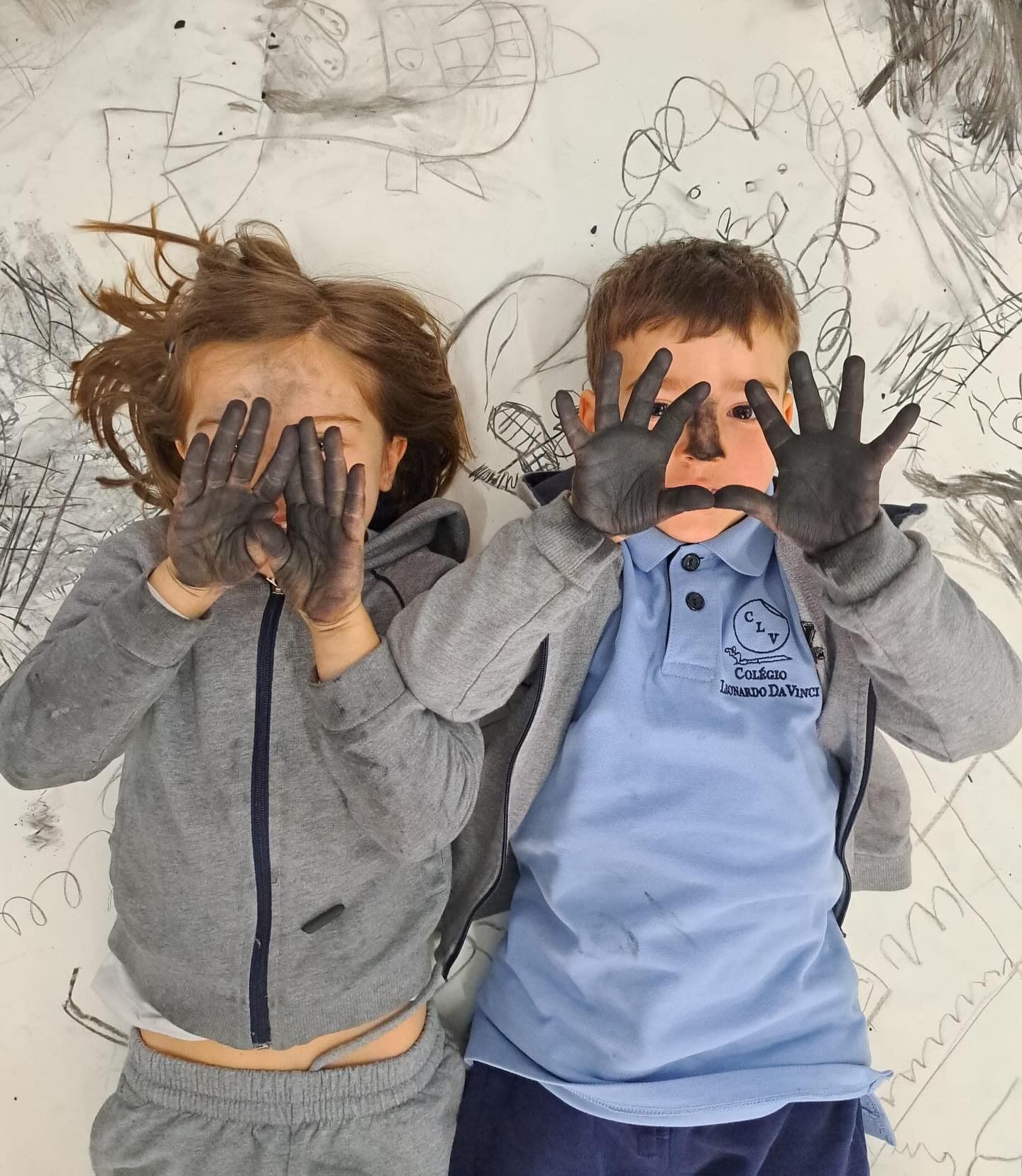 Dois alunos deitados sobre desenhos a carvão, com as mãos pintadas a tapar a cara